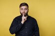 © bodnarphoto - Man with a playful expression holds a finger to his lips against a bright yellow background in a moment of quiet fun