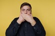 © bodnarphoto - Surprised man covers mouth in disbelief against bright yellow backdrop