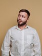 © bodnarphoto - Young man with a beard in a white shirt gazes thoughtfully against a warm beige background