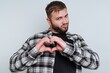 © bodnarphoto - Man wears plaid shirt, shows love with heart gesture against simple backdrop in cozy indoor setting