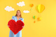 © Pixel-Shot - Young woman with heart near decorated yellow wall. Valentine's Day celebration
