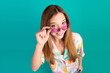 © deagreez - Young Woman in Colorful Shirt Adjusting Pink Sunglasses Against a Teal Background