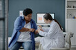© Seventyfour - Middle aged Black woman receiving dermatological examination from young adult Caucasian female doctor applying lotion to arm, visiting medical office with computer monitor in background