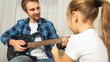 © Mariia - Dad teaches daughter to play guitar in living room. Dad and daughter spend time together.