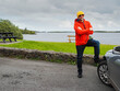 © mark_gusev - A tourist man in red jacket is standing by a car with picturesque lake in the background. The sky is cloudy and the water is calm. Travel and tourism. Calm and relaxing mood.