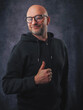© mark_gusev - A man wearing a black hoodie and glasses is smiling. He is standing in front of a grey background. Classic portrait of a confident and friendly looking male model. Thumbs up gesture.