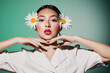 © SHOTPRIME STUDIO - Young woman with bright makeup and two daisies in her hair poses with hands folded under her chin on a pastel green background. Creative concept in beauty fashion style.