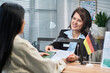 © pressmaster - Caucasian middle aged woman sitting at visa center desk assisting young adult woman with application documents, both interacting across glass partition, German flag on counter