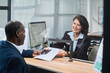 © pressmaster - Middle aged Caucasian woman smiling while assisting middle aged Black man submitting documents at visa center reception desk, both engaging in formal application process through glass partition