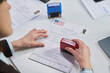 © pressmaster - Caucasian middle aged woman stamping visa application form with rejected stamp at visa center, visible hands holding document, official paperwork and approval stamp on desk