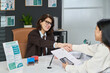 © pressmaster - Caucasian middle aged woman sitting at desk shaking hands with young adult Latin woman, while reviewing visa application documents in modern visa center office setting