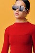 © SHOTPRIME STUDIO - Woman with sunglasses and red sweater posing in studio on yellow background. Fashionable female model wearing trendy eyewear and elegant casual outfit with neutral expression.