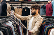© standret - Male shopper examining a checkered shirt while browsing clothing racks filled with various garments in a retail store setting