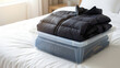 © Guillotine - Winter clothes neatly folded and stored in a clear plastic box on a white bed in a bright bedroom.