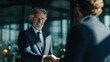 © Jam - Two businessmen shake hands over a desk in a modern office. One man is older with gray hair, while the other is younger and not fully visible