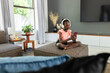 © Wavebreak Media - Teenage female sitting on gray ottoman at home, wearing headphones and holding pink smartphone