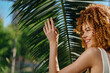 © SHOTPRIME STUDIO - smiling woman with curly hair enjoys tropical sunlight near palm leaves, showing natural beauty and warmth in a bright outdoor setting