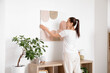 © Pixel-Shot - Young beautiful woman hanging picture on white wall in stylish room