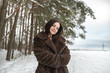 © shymar27 - Female model wearing a brown fur coat and purple skirt stands in a snowy forest, snowflakes falling gently around her, with trees in the background