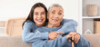 © Pixel-Shot - Happy young woman hugging her grandmother with stick sitting on sofa at home