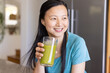 © Wavebreak Media - Asian woman in teal shirt sitting at kitchen counter holding tall glass of green smoothie smiling
