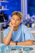 © Nataliia - Young male with curly hair resting chin on hand, wearing blue t-shirt, seated at a table with tools and materials in a bright workshop environment