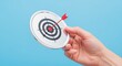 © The Little Hut - Conceptual Image of Hand Holding Target with Arrow Hitting Bulls-eye on Light Blue Background