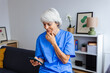 © Xavier Lorenzo - Portrait of senior healthcare worker in blue scrubs relaxing on sofa using mobile phone app or reading an e-mail after workday at hospital. Technology and healthcare business concept