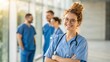 © Alam - Closeup portrait of a smiling nurse with stethoscope and arms folded in a hospital corridor, with her medical team behind
