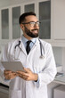 © fizkes - Inspired by medical innovations. Cheerful young male doctor healthcare professional in lab coat hold touchscreen pad in clinic office look away enjoy modern digital tools for patient care improvement