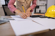 © SOMKID - Close-up of female architect or civil engineer hand drawing blueprint with pencil on workspace table, featuring yellow safety helmet and calculator. Professional construction planning and interior des