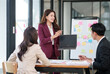 © aekachai - Group of young Asian professionals engaging in a dynamic brainstorming session. A female leader stands confidently while presenting ideas to her colleagues, showcasing a collaborative, positive