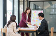© aekachai - Group of young Asian professionals engaging in a dynamic brainstorming session. A female leader stands confidently while presenting ideas to her colleagues, showcasing a collaborative, positive