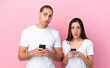 © luismolinero - Young caucasian couple isolated on pink background looking at the camera while using the mobile with surprised expression
