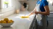 © ALLmost - A person in an apron cleans a white countertop with a spray bottle and yellow cloth in a sunlit kitchen.