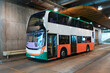 © aapsky - A contemporary double-decker bus in white, orange, and green sits stationary within a bright indoor public transport facility. Concrete walls and ceiling lights illuminate the spacious depot
