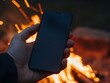 © motiva_dsgn - Hand holding smartphone with blank screen near campfire at night