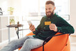 © Prostock-studio - Mobile Shopping. Cheerful Man Using Phone And Credit Card Purchasing Things In Application Sitting In Chair At Home. Side View Of Guy Holding Bank Card And Smartphone Using Bank App Online