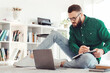 © Prostock-studio - Millennial Guy Writing Taking Notes Looking At Laptop Computer Making Business Report Working Online Sitting On Floor At Home, Wearing Eyewear. Freelance Concept. Selective Focus