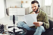 © Prostock-studio - Man Reading Papers Video Calling Via Smartphone On Tripod And Wearing Headset Sitting In Chair At Home. Modern Business Communication And Remote Work Concept. Selective Focus