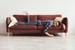 © Prostock-studio - A man is lying on a couch in a relaxed position with one leg hanging off the side and arms stretched out. He appears to be taking a break in a living room.
