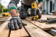 © SKIMP Art - a worker is working on the wooden floor of an outdoor area he has on black gloves and a gray shirt with green accents in his hand, there's a yellow drill and other tools Generative AI
