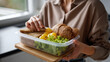 © Taskmanager - Woman Holding Healthy Lunch Box with Fresh Greens, Eggs, Whole Grain Bread, and Baked Goods in Modern Kitchen Setting