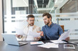 © Liubomir - Two male business professionals are reviewing documents and discussing a project, collaborating effectively at a conference table with laptops and papers in a contemporary corporate office setting