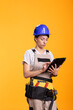 © DC Studio - Caucasian female contractor checks digital plans on tablet with focused expression, symbolizing planning or project review. Woman renovator wearing tool belt stands in studio against yellow backdrop.
