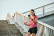 © Stockphotodirectors - A woman in athletic wear stands on a staircase, smiling as she listens to music on her headphones and checks her phone while holding a water bottle, promoting a healthy lifestyle outdoors.
