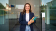 © Miquel - Confident young businesswoman holding folders smiling at office entrance