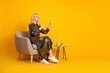 © deagreez - Glamorous mature woman sits in a comfy armchair wearing a patterned dress smiling and pointing with both fingers in a bright yellow studio