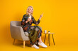 © deagreez - Elegant mature woman sits in a beige chair against a bright yellow backdrop holding a phone and pointing up in a stylish fashion photo