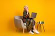 © deagreez - Elegant mature woman in a floral dress sits in a beige chair with laptop on a yellow studio background smiling and thinking about work and style
